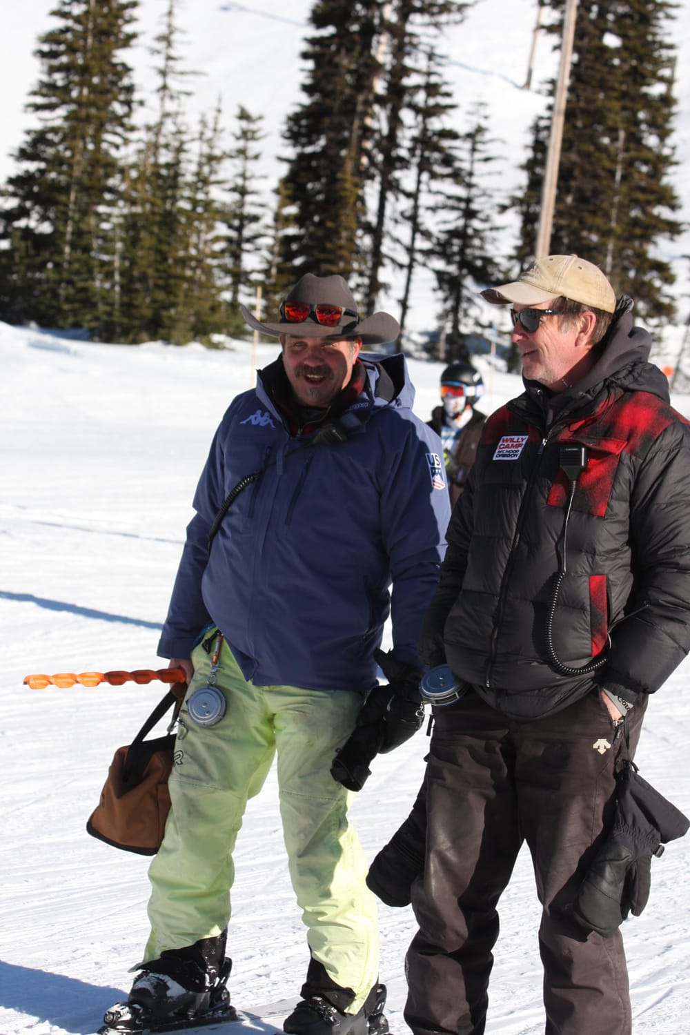 Two coaches talking on the hill, one in a Willy Camp jacket with a radio clipped to the chest